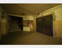 Ein unterirdischer Bunkerraum mit Betonwänden und -decke, einer Treppe links, einem Registraturbereich mit hölzernem Schaukasten in der Mitte und einer massiven schwarzen Stahltür rechts. Die Beleuchtung ist schwach, und die Umgebung wirkt funktional und schlicht.