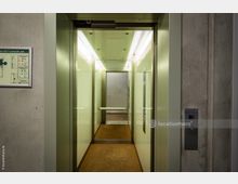 Ein moderner Fahrstuhl mit offenstehenden Türen; der Innenraum ist hell beleuchtet, mit beigen Wänden, Spiegeln und einem Handlauf. Links ist ein Evakuierungsplan an der Betonwand angebracht, rechts ein silbernes Bedienfeld mit einem roten Licht.
