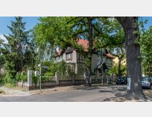 Straßenszene mit einem großen, historischen Wohnhaus im Fachwerkstil, umgeben von Bäumen und einer Ziegelmauer mit Zaun. Im Vordergrund ist eine Straße mit parkenden Autos und ein Straßenschild zu sehen.
