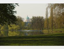 Weitläufiger Park mit Grünflächen, Seen und Bäumen im Vordergrund, im Hintergrund eine Kuppel eines historischen Gebäudes, die über die Baumkronen hinweg sichtbar ist.