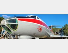 Nahaufnahme eines silberfarbenen Flugzeugs mit rotem Streifen und historischem Logo an der Seite, geparkt in einer Außenumgebung mit blauem Himmel und Bäumen im Hintergrund. Einblick in die Cockpitfenster und Details des vorderen Bereichs des Flugzeugs sichtbar.