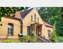 Backsteingebäude mit gelben und roten Klinkersteinen, Satteldach und großen Fenstern mit Rundbögen. Vor dem Gebäude führt eine Treppe zur Eingangstür, umgeben von Bäumen und grüner Vegetation.