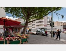 Straßenansicht mit einem Obststand unter einem Baum im Vordergrund, der Melonen und andere Früchte anbietet. Im Hintergrund sind Wohn- und Geschäftsgebäude, parkende Autos, ein Bus und Passanten an einer Ampel zu sehen.