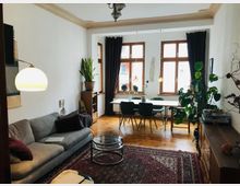 Helles Wohnzimmer mit Holzboden, einer grauen Couch und einem Glastisch im Vordergrund. Im Hintergrund ein Essbereich mit Holzstühlen, großen Fenstern mit Vorhängen und mehreren Zimmerpflanzen.