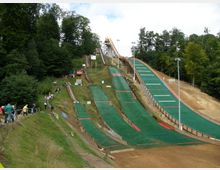 Skisprungschanze in einer waldreichen Umgebung mit mehreren mit grünem Kunststoff bedeckten Schanzenanlagen, umgeben von Zuschauern auf einem Grasweg. Im Hintergrund sind Bäume und eine größere Schanze sichtbar.