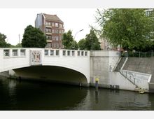 Weiße Brücke mit dekorativen Gittern, die über einen Fluss führt, daneben eine betonierte Treppe mit Graffiti. Im Hintergrund steht ein rotes Backsteingebäude und mehrere Bäume.