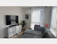 Helles, modern eingerichtetes Wohnzimmer mit großem grauen Ecksofa, wandmontiertem Flachbildfernseher über einem weißen Sideboard und einem runden Hängelampenschirm. Große Fenster mit transparenten Vorhängen lassen viel Tageslicht herein; eine Zimmerpflanze, ein gerahmtes Bild und eine kleine Tischlampe setzen Akzente.