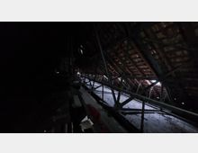 Dunkler Dachboden mit sichtbarer Holz- und Metallkonstruktion, sowie teilweise durchlässigem Ziegeldach, durch das Lichtstrahlen einfallen. Der Boden ist mit Staub oder Schutt bedeckt, und die Umgebung wirkt unfertig oder ungenutzt.