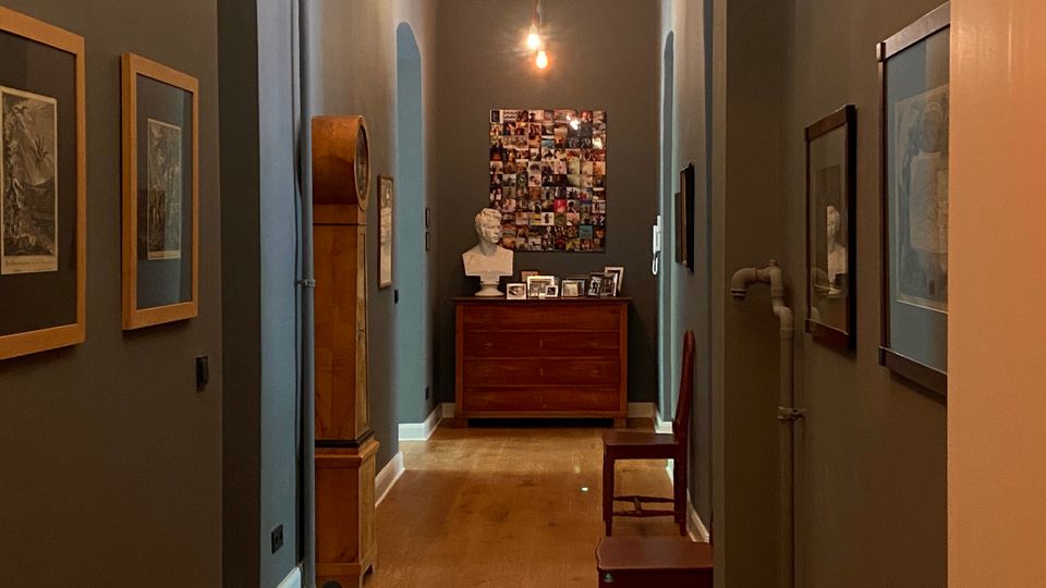 Langer Innenflur mit dunklen Wänden, Holzboden und gewölbter Decke, beleuchtet durch mehrere hängende Glühbirnen. An den Wänden hängen gerahmte Bilder; am Ende stehen eine Kommode mit Foto-Collage darüber sowie ein hoher Standuhrschrank und zwei Stühle.
