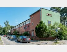 Eine Reihe von mehrfarbigen Reihenhäusern mit Flachdächern in einem grünen Wohngebiet, umgeben von Bäumen und Sträuchern. Entlang der gepflasterten Straße stehen geparkte Autos, im Vordergrund befindet sich ein Straßenschild.