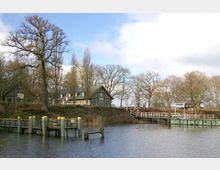 Ruhige Uferlandschaft mit Anlegestelle und Gebäuden: Im Vordergrund ein kleiner Steg mit gelben und blauen Pfosten am Ufer eines Gewässers. Dahinter stehen mehrere traditionelle Häuser und Bäume, teilweise ohne Laub, unter einem bewölkten Himmel. Ein Schild mit der Aufschrift „Pfaueninsel“ ist sichtbar.