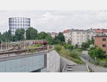 Eine Stadtansicht mit einer vorbeifahrenden S-Bahn auf erhöhten Gleisen, flankiert von Bäumen und mehrstöckigen Wohngebäuden im Hintergrund. Links ist ein großer Gasometer zu sehen, rechts befindet sich eine niedrige Betonmauer und ein modern verglastes Gebäude.
