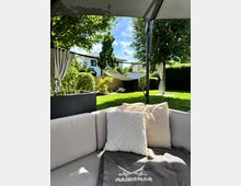 Terrasse mit Sitzbereich und hellen Kissen im Vordergrund, umgeben von einem gepflegten Garten mit Rasen, Bäumen und einer aufgehängten Hängematte. Im Hintergrund sind Hecken und moderne Häuser zu sehen.