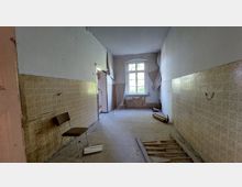 Ein schmaler, leerer Flur in einem verfallenen Gebäude mit abblätternder Tapete, einer großen Fensterfront am Ende und vergilbten Fliesen an den Wänden. Auf dem Boden liegen Holzstücke, und links steht ein einzelner alter Stuhl.