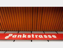 Rotes Stationsschild mit der Aufschrift „Pankstraße“ in weißer Schrift, vor einer Decke aus vertikalen, orangefarbenen Holzpaneelen. Darunter sind graue, rechteckige Wandplatten sichtbar.