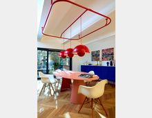 Moderner Essbereich mit einem ovalen, rosafarbenen Tisch, umgeben von weißen Stühlen mit Holzbeinen. Rote, kugelförmige Pendelleuchten hängen von einer roten Metallstruktur an der Decke, während im Hintergrund ein blaues Sideboard und großflächige Kunstwerke zu sehen sind; Tageslicht fällt durch große Glasfenster auf den Fischgrät-Holzboden.