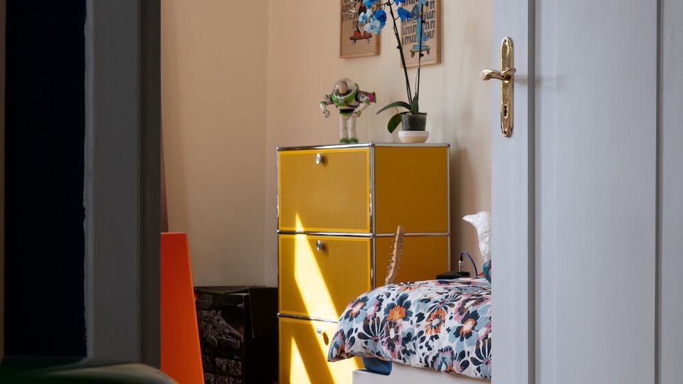 View through a partially open white door into a small bedroom with cream walls, a yellow modular dresser, and a white bed with a floral duvet. Sunlight falls across the wooden floor, and framed wall art and a potted orchid sit above the dresser.