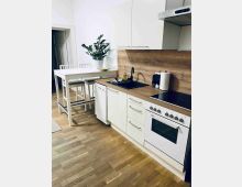 Helle, moderne Küche mit weißen Schränken, Holzarbeitsplatte und durchgehender Holzrückwand. Sichtbar sind Spüle mit schwarzer Armatur, Herd mit Backofen und Dunstabzugshaube sowie ein schmaler Esstisch mit hohen Stühlen auf Holzboden.