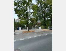 Straßenansicht mit einer gepflasterten Gehwegfläche, mehreren Bäumen und einem umzäunten Grundstück mit einem weißen Einfamilienhaus im Hintergrund. Das Haus ist teilweise verdeckt von Bäumen und Sträuchern, und am Straßenrand stehen Autos.