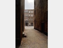 Schmale Gasse mit Kopfsteinpflaster, flankiert von hohen Backsteinmauern, die zu einem offenen Hof mit einem mehrstöckigen historischen Gebäude aus Ziegelstein führen. Im Hintergrund sind große Fenster und Eingangstüren sichtbar.