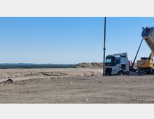 Eine Baustelle mit einer großen, kahlen Erdfläche, einem weißen Lkw und einem gelben Bagger der Marke CAT. Im Hintergrund sind Erdhügel und eine grüne Waldlandschaft unter klarem, blauem Himmel sichtbar.