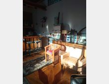 Ein gemütlicher Innenraum mit Holzboden und moderner Einrichtung, darunter ein hölzerner Stuhl, ein beiger Sessel und eine Theke mit Barhockern. Im Hintergrund stehen Kunstwerke, Staffeleien und Dekorationsobjekte, während Sonnenlicht durch ein Fenster den Raum beleuchtet.