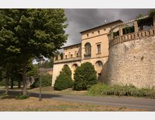 Historisches Gebäude mit gelber Fassade und Rundbogenfenstern, flankiert von einer Steinmauer mit Pergola-Struktur; im Vordergrund Bäume, eine Laterne und ein Gehweg.