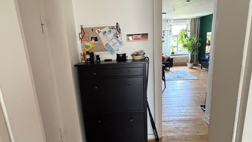 Schmaler Flur mit weißen Wänden und Holzdielenboden, links eine schwarze Schuhkommode mit Pinnwand und kleiner Dekoration. Durch eine offene Tür blickt man in ein helles Wohnzimmer mit großem Fenster, Teppich und einer Zimmerpflanze.