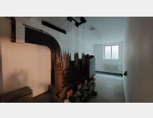 Ein technischer Raum mit weiß gestrichenen Wänden, einer großen mechanischen Anlage im Vordergrund und einem rechteckigen Fenster im Hintergrund, das Tageslicht hereinlässt. An der rechten Wand befindet sich ein Heizkörper unter dem Fenster.