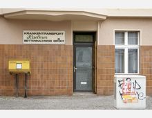 Fassade eines Gebäudes mit braunen Fliesen; links ein gelber Briefkasten, in der Mitte eine Glastür, rechts ein Fenster. Ein weißer Kasten mit Plakaten und Graffiti steht rechts auf dem Gehweg. Ein Schild über der Tür zeigt: "Krankentransport Haeberer".