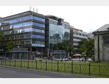 Moderne Bürogebäude mit gläserner Fassade und angrenzenden Gebäuden aus verschiedenen Baustilen umgeben eine kleine Grünfläche mit Gehwegen. Im Vordergrund befindet sich ein Metallzaun, und Passanten beleben den Bereich.