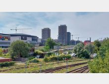 Blick auf eine Stadtlandschaft mit Bahngleisen im Vordergrund, dichter Vegetation und kleinen Gebäuden. Im Hintergrund sind die Mercedes-Benz Arena, im Bau befindliche Hochhäuser mit Baukränen sowie weitere Gewerbebauten zu sehen.