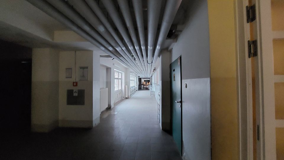 Langer, schmaler Flur in einem Gebäude mit weißen Wänden und dunklem Boden, über dem zahlreiche Rohre an der Decke verlaufen; auf der rechten Seite eine geschlossene grüne Tür und auf der linken Seite Fenster, die diffuses Tageslicht hereinlassen.