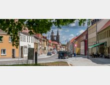Straßenansicht einer kleinen Stadt mit bunt gestrichenen Häuserfassaden, einem Café mit Außensitzplätzen und geparkten Autos am Straßenrand. Im Hintergrund ragt eine große Kirche mit zwei Türmen und Spitzen hervor, umgeben von einem bewölkten Himmel.