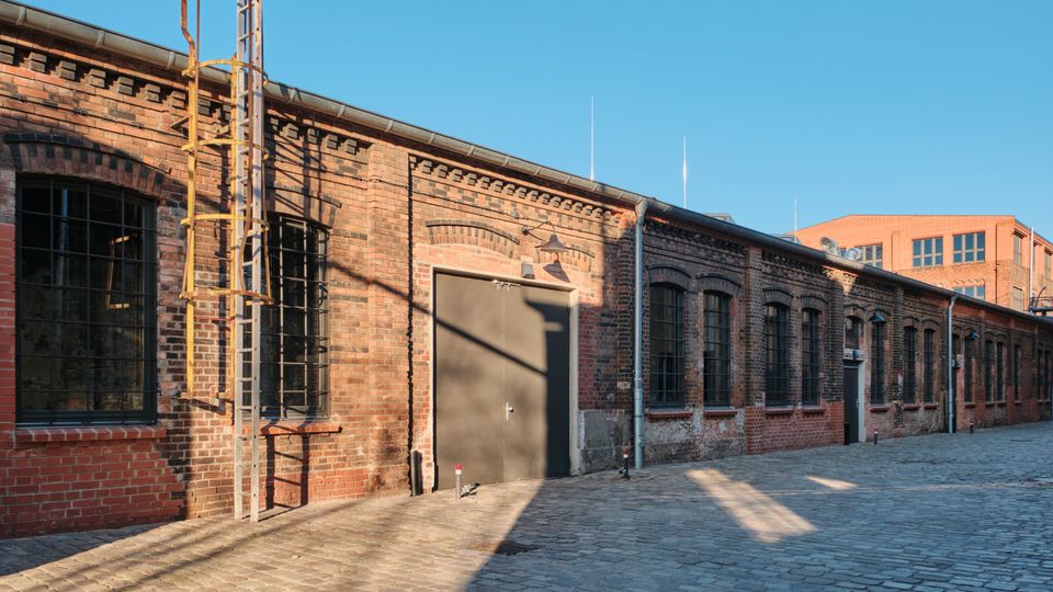 Long red-brick industrial building with arched, metal-barred windows and a large gray door, bordering a cobblestone courtyard. A vertical metal ladder and rooftop gutters line the facade under a clear blue sky, with another brick building visible in the background.