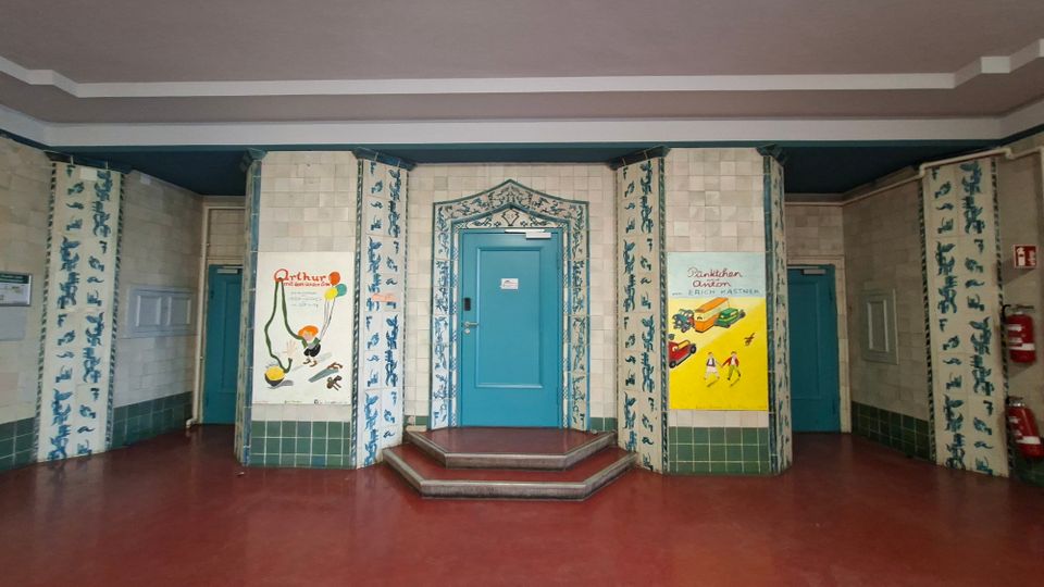 Innenraum eines gefliesten Treppenhaus- oder Flurbereichs mit rotbraunem Boden und mehreren türkisfarbenen Türen, die von blau gemusterten Fliesenbändern gerahmt sind. In der Mitte führt eine kurze, gestufte Plattform zu einer zentralen Tür; an den Wänden hängen farbige Plakate und rechts sind Feuerlöscher angebracht.