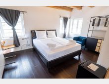 Helles Schlafzimmer mit Doppelbett, dunklem Holzfußboden und sichtbaren Deckenbalken. Drei Fenster mit grauen Vorhängen, daneben ein blaues Sofa und ein offenes Regal- und Schrankelement an der Wand.