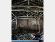 Innenansicht einer alten Scheune mit Holzbalken und Ziegeldach. Der Boden ist mit Heu und verstreutem Stroh bedeckt, an der Ziegelwand lehnen einige ältere Gegenstände, darunter ein rotes Möbelstück.