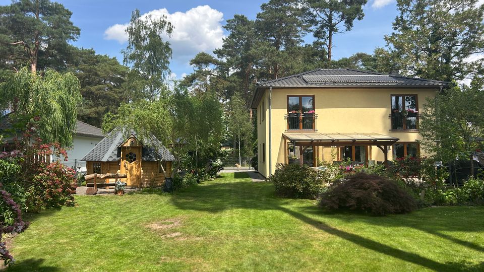Großer, gepflegter Garten mit Rasenfläche, umgeben von Sträuchern und hohen Bäumen, mit einem zweistöckigen Wohnhaus mit Terrasse sowie einem kleinen Holz-Gartenhaus mit Schindeldach. Im Hintergrund sind weitere Häuser und ein blauer Himmel mit Wolken zu sehen.