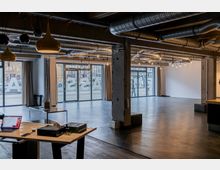 Großer, moderner Innenraum mit industriellem Design, sichtbar durch freiliegende Lüftungsrohre und Betonpfeiler. Glasfassaden mit großen Fenstern bieten Blick auf ein städtisches Umfeld; der Raum enthält vereinzelte Möbel wie Tische und Hocker.