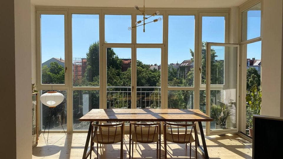 Bright dining area with a wooden table and chairs set in front of large floor-to-ceiling windows and a glass door opening to a small balcony. White-framed windows fill the wall, letting in sunlight and views of trees and nearby rooftops, with a modern ceiling light fixture overhead.