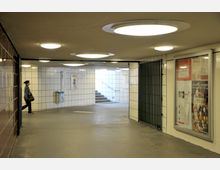 Unterführung mit weiß gekachelten Wänden, abgerundet durch runde Deckenleuchten. Auf der rechten Seite befinden sich Informationsplakate hinter Glas, links läuft eine Person in Richtung Ausgang mit einer Treppe im Hintergrund.