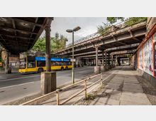 Eine städtische Straßenszene unter einer alten, rostigen Eisenbahnüberführung mit filigranen Pfeilern aus Metall. Rechts neben der Straße verläuft ein Gehweg mit Plakaten an der Wand, während ein gelber Doppeldeckerbus die Fahrbahn überquert.