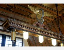 Detailansicht einer hölzernen, bemalten Decke mit kunstvollen Mustern und Verzierungen in warmen Brauntönen. Darunter hängt eine dekorative Leuchte mit drei Glaslampen und einem verzierten Element mit einem goldenen Flügelmotiv.