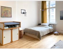 Helles Schlafzimmer mit Holzfußboden, einem Einzelbett neben einem großen Fenster mit goldenen Vorhängen und einer Kommode mit Wickelauflage. An der weißen Wand hängen ein abstraktes Bild und ein kleiner Basketballkorb; daneben stehen Körbe und ein kleines Babybettchen am Boden.