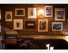 Gemütlicher Innenraum mit einer dunkelroten, goldgemusterten Tapete und gerahmten Schwarz-Weiß-Bildern an der Wand. Im Vordergrund ein dunkelbraunes Ledersofa, ein Holztisch mit einer alten Radiokonsole und brennenden Kerzen.