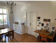 Ein heller Raum mit Holzboden, einer weißen Doppelflügeltür und einem großen Fenster mit langen, weißen Vorhängen. Links steht ein Esstisch mit Stühlen, in der Mitte ein weißer Schrank mit Bildern und Dekoration, und rechts ein Schreibtisch mit Kindermalereien und Schreibmaterialien an der Wand.