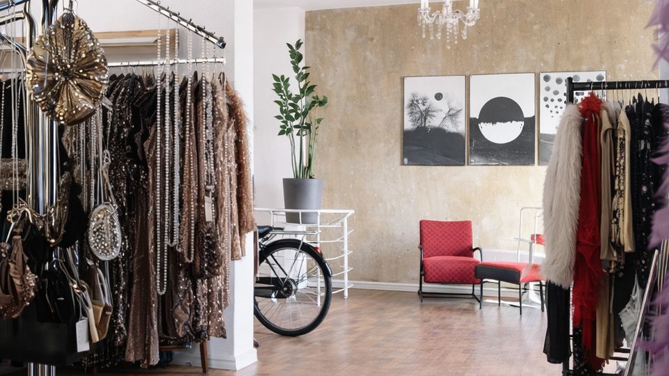Heller, loftartiger Boutique- oder Showroomraum mit Holzboden, Kleiderständern voller glitzernder Kleidung und Taschen sowie Kronleuchtern an der Decke. Im hinteren Bereich stehen rote Sessel vor einer beige strukturierten Wand mit drei gerahmten Bildern; daneben ein Fahrrad und eine Topfpflanze.