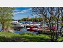 Hafen mit kleinen Booten und Yachten, umgeben von Bäumen und Grünflächen. Im Hintergrund ist eine Kirche mit einem spitzen Turm sichtbar, daneben Gebäude und eine grüne Landschaft.