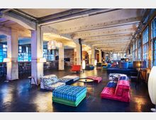 Industrie-Loft mit modernem Interieur, bestehend aus bunten Polstermöbeln, niedrigen Tischen und Stehlampen. Der Raum zeichnet sich durch freiliegende Betonpfeiler, eine hohe Decke mit sichtbaren Trägern und große Fensterfronten aus, die Tageslicht hereinlassen.